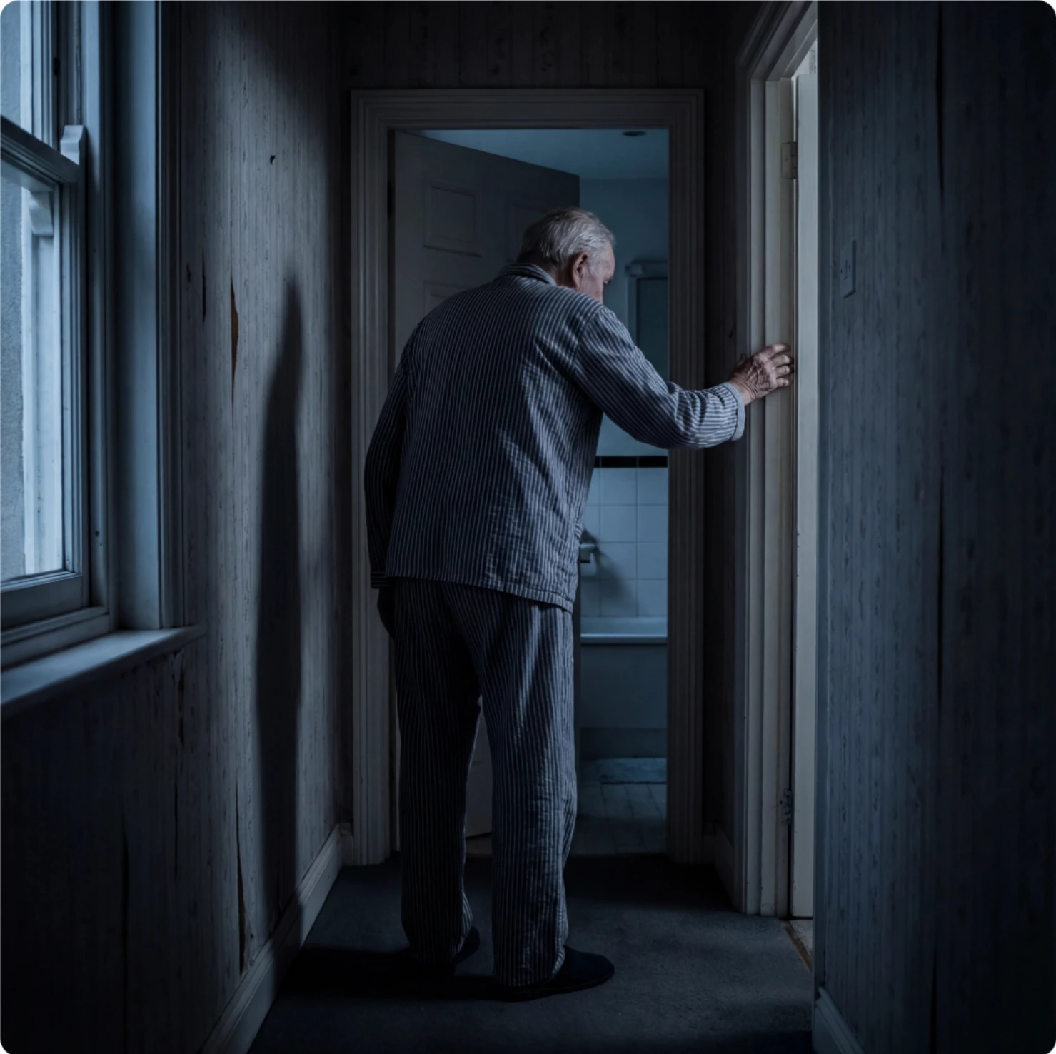 Un homme âgé en pyjama, la main appuyée sur le montant de la porte, traverse un couloir sombre vers la salle de bain la nuit.